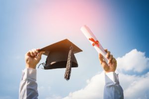 A person celebrating their new academic achievement by holding a graduation cap and diploma in the air.