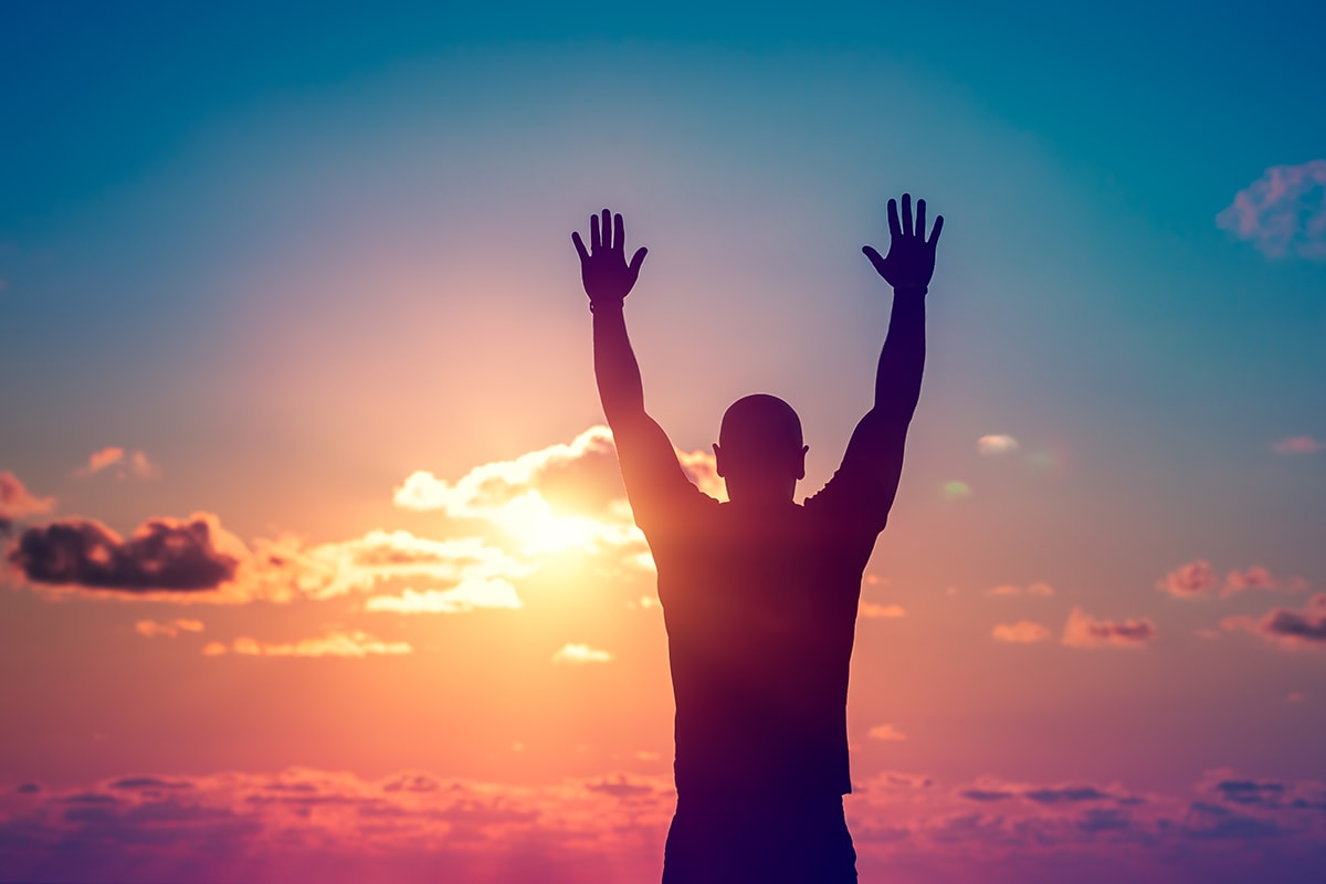 اكتساب الثقة بالنفس- A man's silhouette with hands raised at sunset.